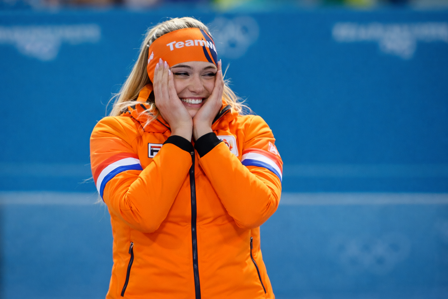 Nederlandse schaatsster in oranje TeamNL-jack en hoofdband glimlacht breed en houdt haar handen tegen haar gezicht op een ijsbaan